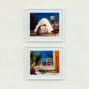 Two white framed photographs by Richard Heeps. The the top photograph, in the foreground of the photograph is a broken off head of a giant fibreglass sculpture of a cowboy (a Muffler Man). In the background set in a deep blue sky is the muffler man's headless torso. The bottom photograph shows a colourful Californian motel against a blue sky.