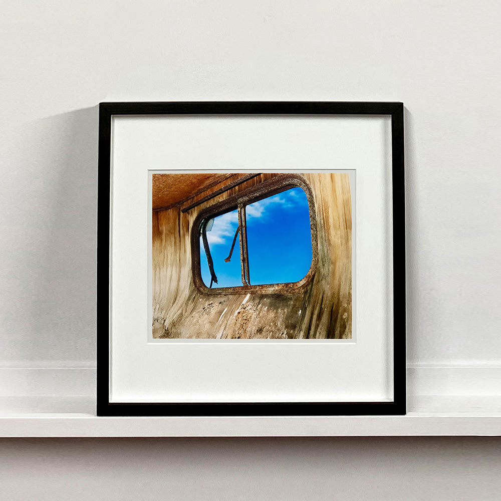 Black framed photograph by Richard Heeps. The interior of an eroded trailer, looking through the broken window to a deep blue sky.