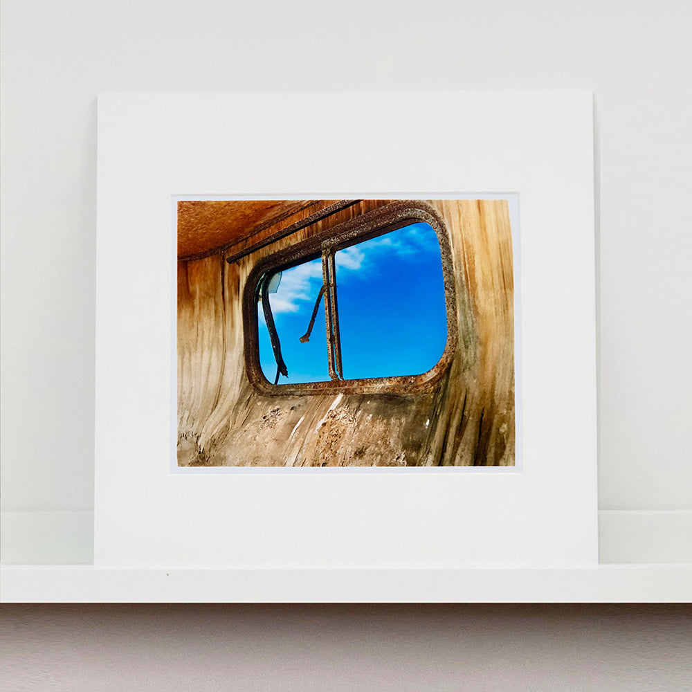 Photograph by Richard Heeps. The interior of an eroded trailer, looking through the broken window to a deep blue sky.