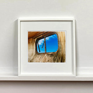 White framed photograph by Richard Heeps. The interior of an eroded trailer, looking through the broken window to a deep blue sky.