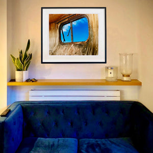 In situ black framed photograph by Richard Heeps. The interior of an eroded trailer, looking through the broken window to a deep blue sky.