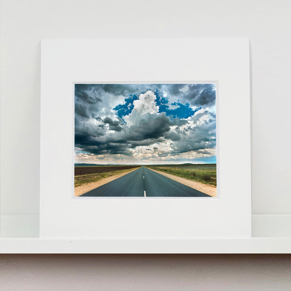 Mounted photograph by Richard Heeps. A photograph looking along a long straight road into the distance, it is edged either side with sand and then prairie land. The vast, cloudy sky sits above.