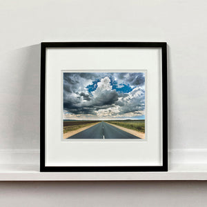 Black framed photograph by Richard Heeps. A photograph looking along a long straight road into the distance, it is edged either side with sand and then prairie land. The vast, cloudy sky sits above.