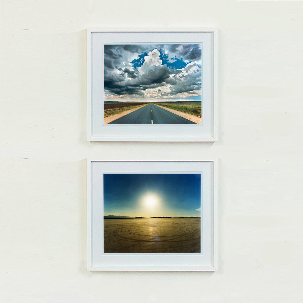 Two white framed photographs by Richard Heeps. The top one is a photograph looking along a long straight road into the distance, it is edged either side with sand and then prairie land. The vast, cloudy sky sits above. The photo below is a vast sky with a hazy sun shining and below is a flat beige ground with circular marks on it.