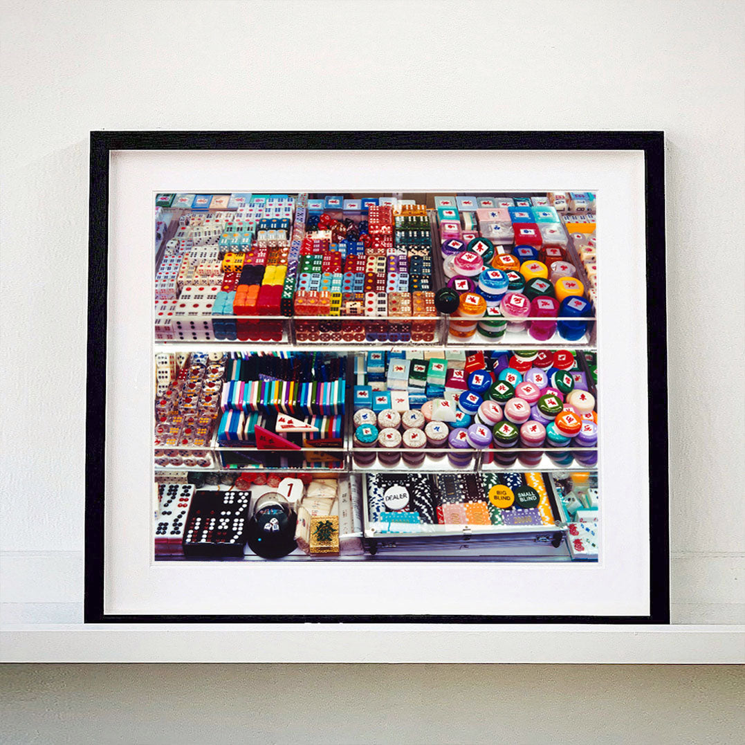 Black framed photograph by Richard Heeps. Three compartmentalised shop shelves, each compartment has coloured plastic dice, counters, circular containers, cards, poker chips, and other plastic type games items.