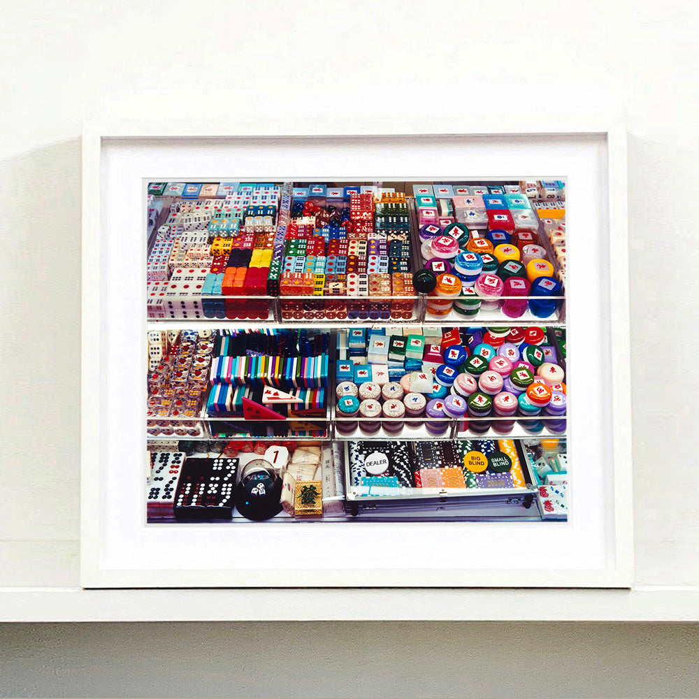 White framed photograph by Richard Heeps. Three compartmentalised shop shelves, each compartment has coloured plastic dice, counters, circular containers, cards, poker chips, and other plastic type games items.