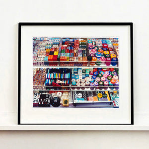 Black framed photograph by Richard Heeps. Three compartmentalised shop shelves, each compartment has coloured plastic dice, counters, circular containers, cards, poker chips, and other plastic type games items.