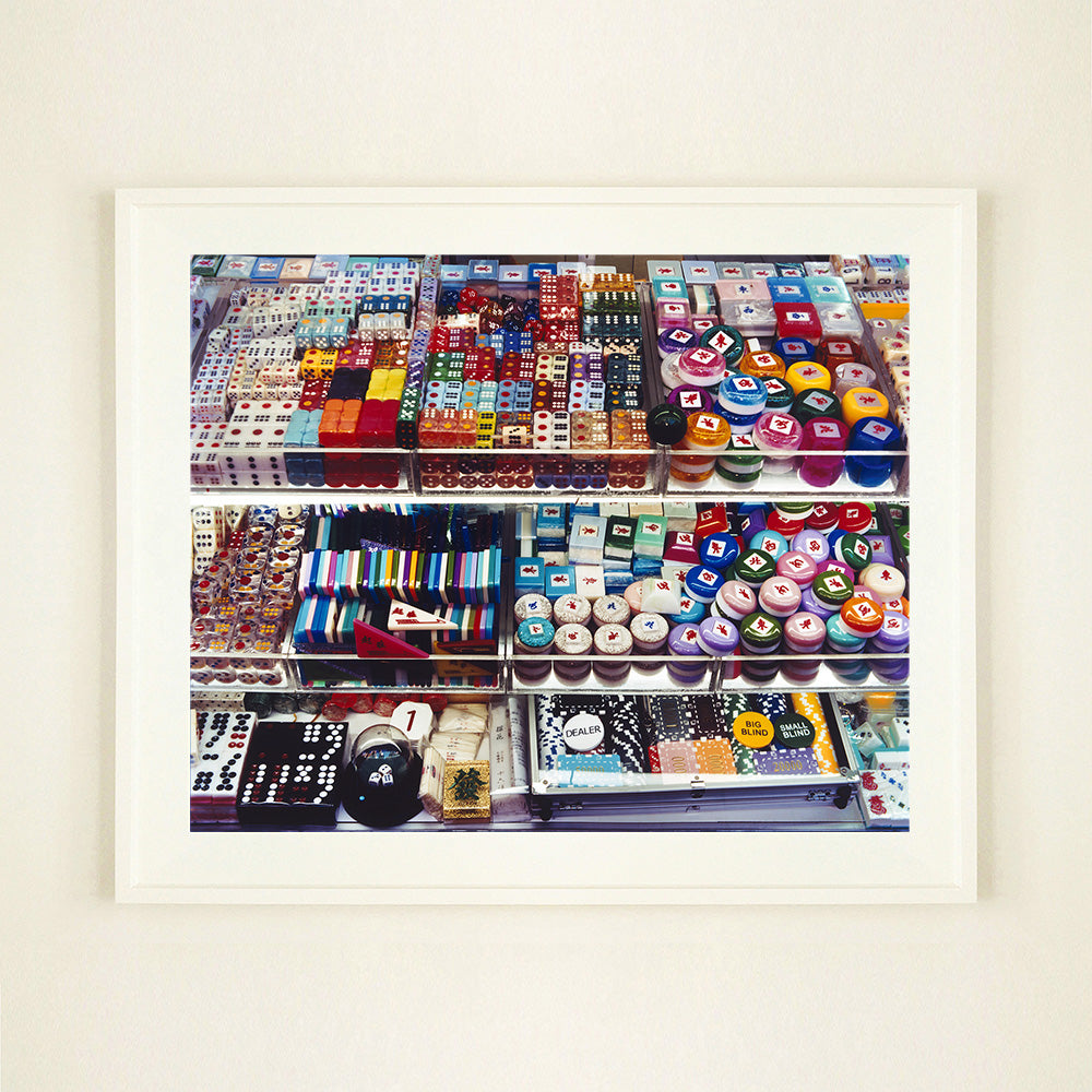 White framed photograph by Richard Heeps. Three compartmentalised shop shelves, each compartment has coloured plastic dice, counters, circular containers, cards, poker chips, and other plastic type games items.