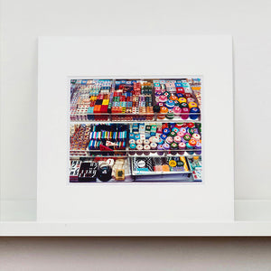 Mounted photograph by Richard Heeps. Three compartmentalised shop shelves, each compartment has coloured plastic dice, counters, circular containers, cards, poker chips, and other plastic type games items.