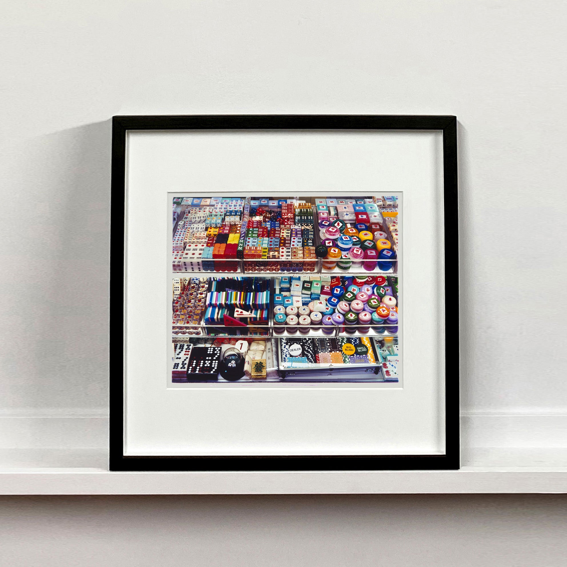 Black framed photograph by Richard Heeps. Three compartmentalised shop shelves, each compartment has coloured plastic dice, counters, circular containers, cards, poker chips, and other plastic type games items.