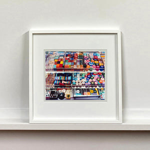 White framed photograph by Richard Heeps. Three compartmentalised shop shelves, each compartment has coloured plastic dice, counters, circular containers, cards, poker chips, and other plastic type games items.