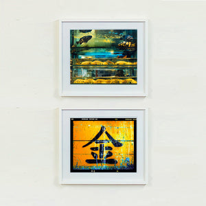 White framed photographs by Richard Heeps. The top photograph shows a wet market fish tank filled with blue green and yellow fish, in a Hong Kong street. The bottom photograph features a black painted Chinese symbol painted on a yellow worn golden canvas.