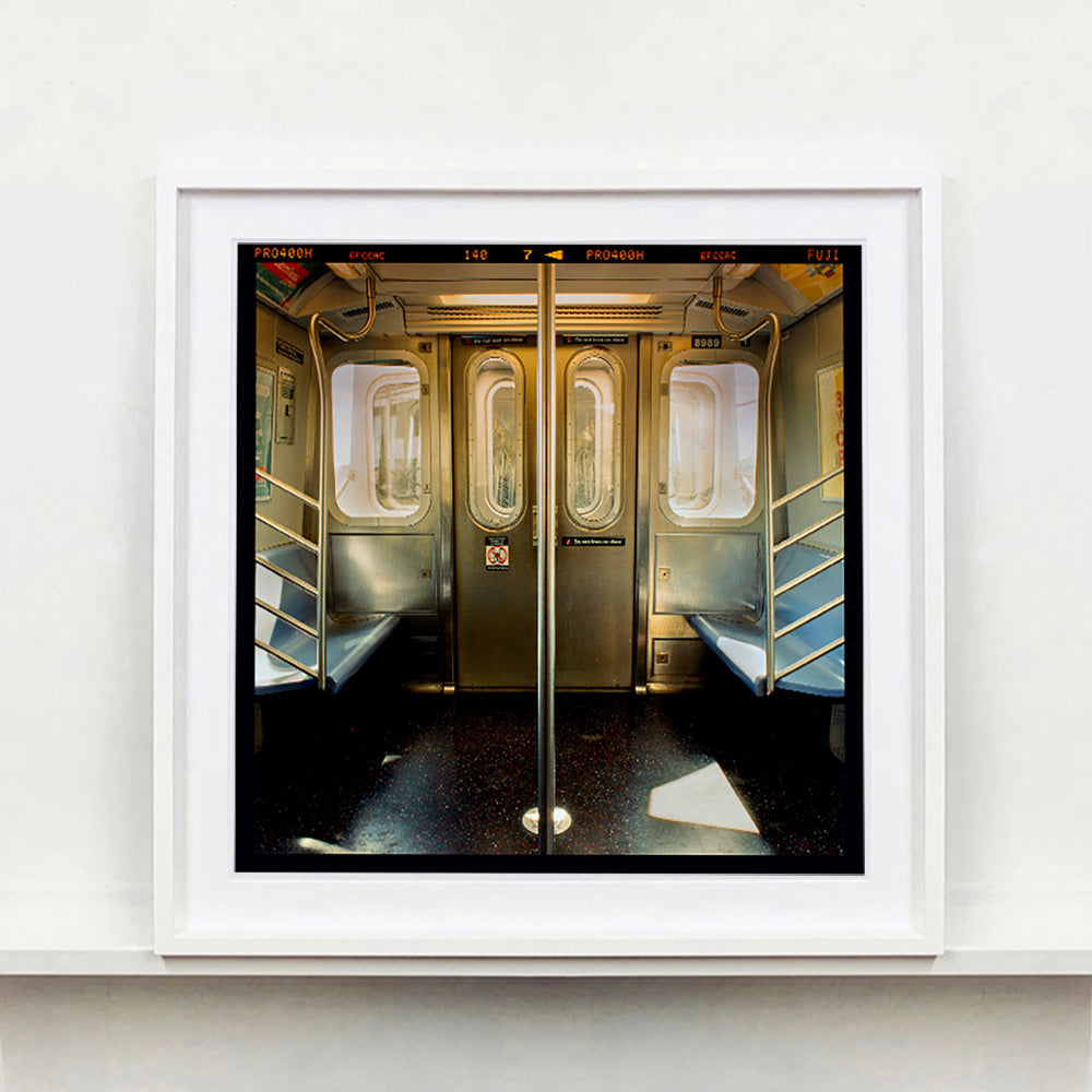 White framed photograph by Richard Heeps. Photograph of the inside of a subway car, looking towards the coaches through the emergency adjoining door. A grab pole sits in the middle.
