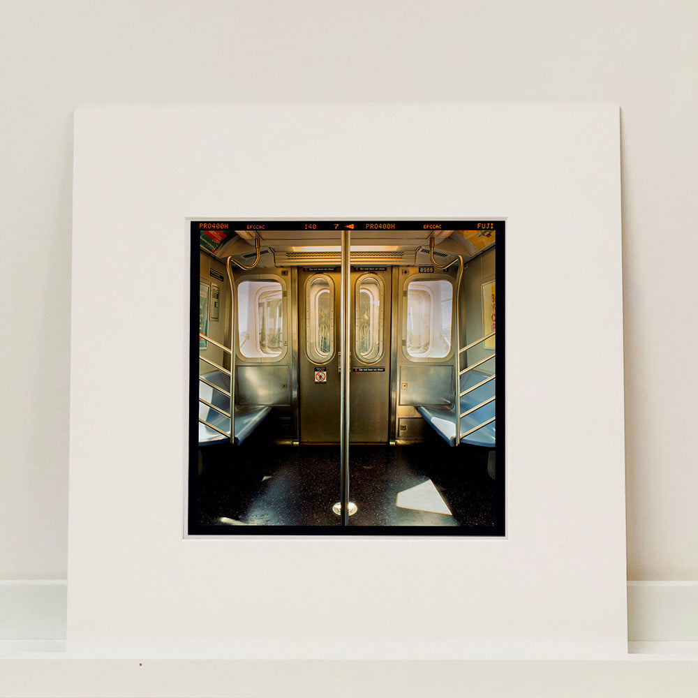 Mounted photograph by Richard Heeps. Photograph of the inside of a subway car, looking towards the coaches through the emergency adjoining door. A grab pole sits in the middle.