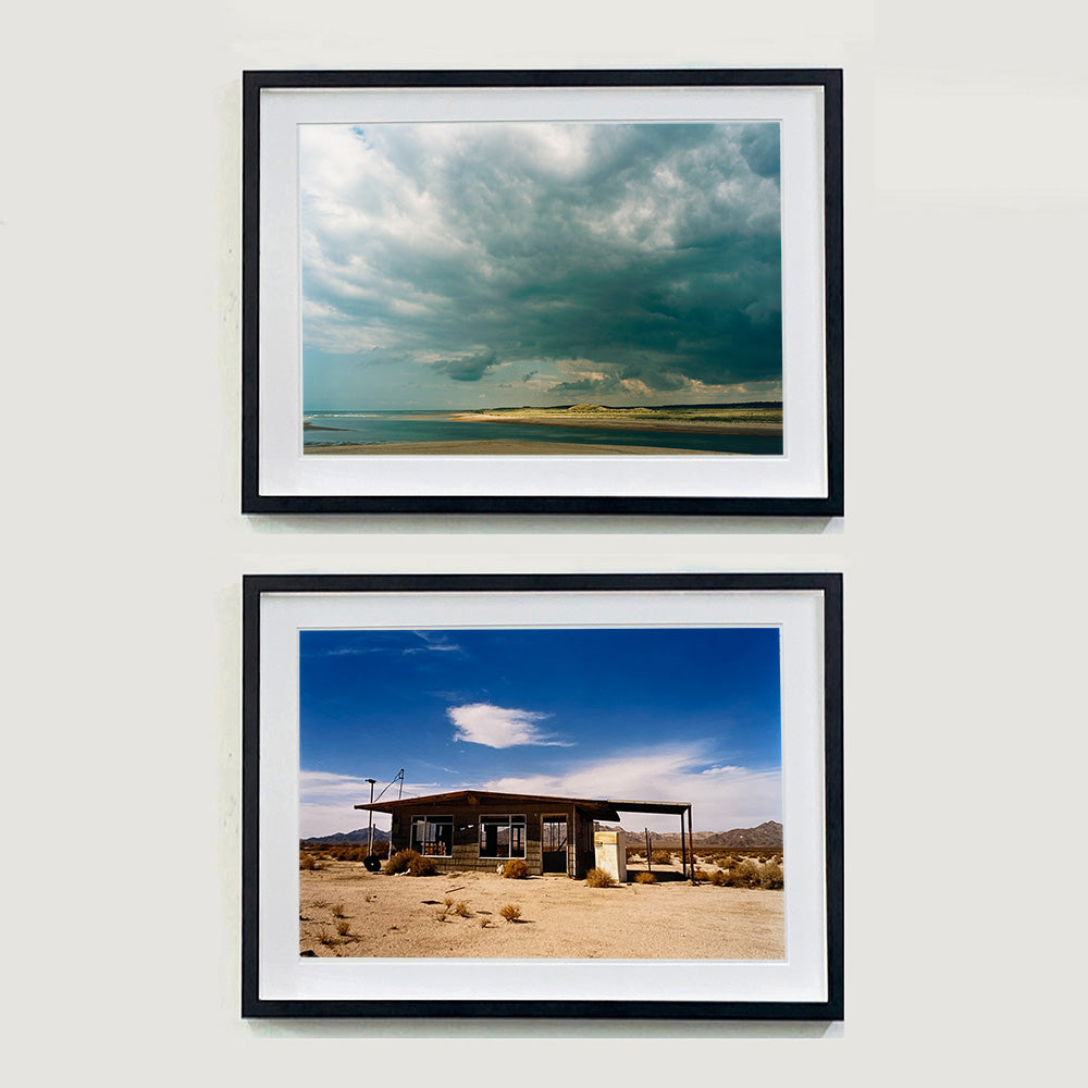 Two black framed photographs by Richard Heeps. The top photograph depicts a dark rolling sky sits over this headland pouring different lights over the sea, sand and dunes below. The bottom photograph is of a derelict shack in the middle of a desert scene, with hills in the distance and overhead a deep blue sky with white fluffy clouds.