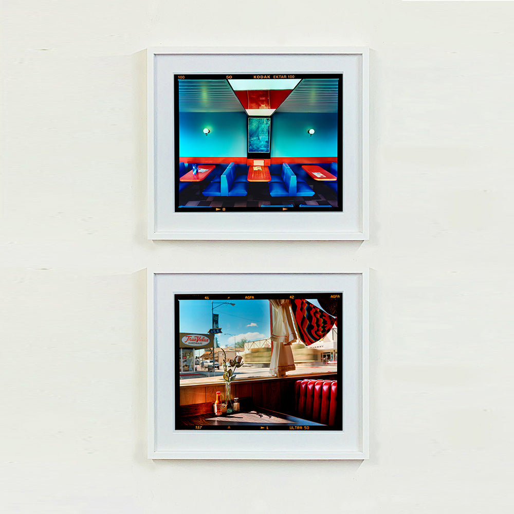 Two white framed photographs by Richard Heeps. The top photograph is of the inside of a Wimpy restaurant. Blue upholstered double seats pinned to the floor and separated by striking red tables. The walls are blue with a blue picture of trees with a black surround aligned with the middle table. The bottom photograph is of the inside of an American diner, with its iconic red upholstered seating, and looking out of the window.