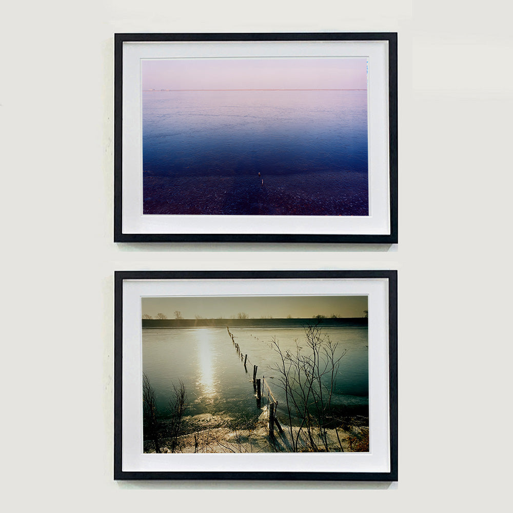 Two black framed photographs by Richard Heeps. The top photograph is looking towards the water. From a distance you can see two blocks of colour, blue at the bottom and lilac at the top, closer up you see the move of the water and a thin land strip along the horizon. The bottom photograph is a watery expanse in neutral tones, a fence cuts vertically through the water and in the distance is a horizontal strip of land.