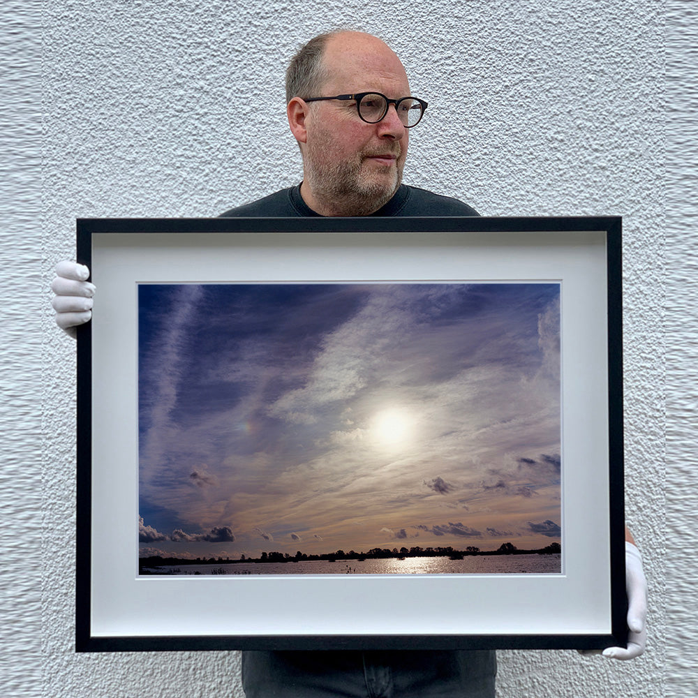 Black framed photograph held by photographer Richard Heeps. A big sky with exciting clouds, an obscured sun and a flood of water features at the bottom reflecting the sun's light.