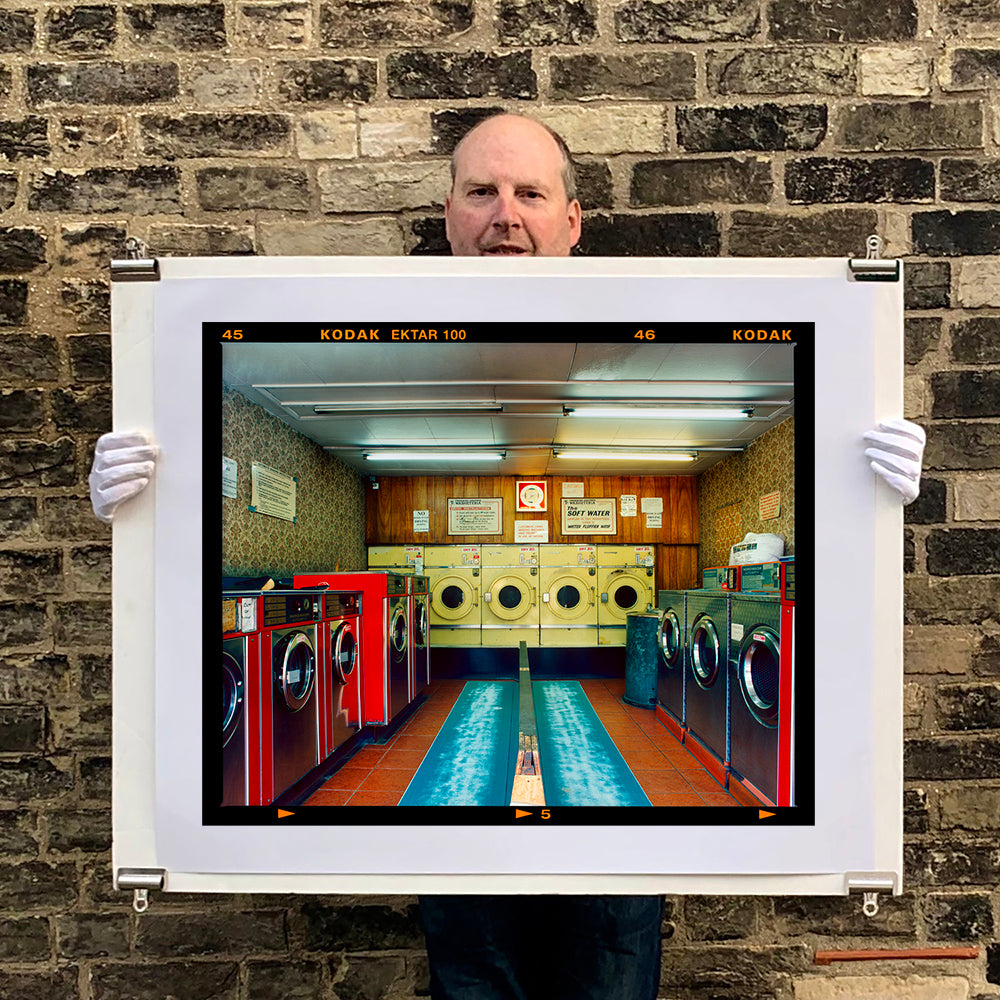 A photograph held by photographer Richard Heeps. A laundrette with washing machines on each wall and a double sided seat in the middle.