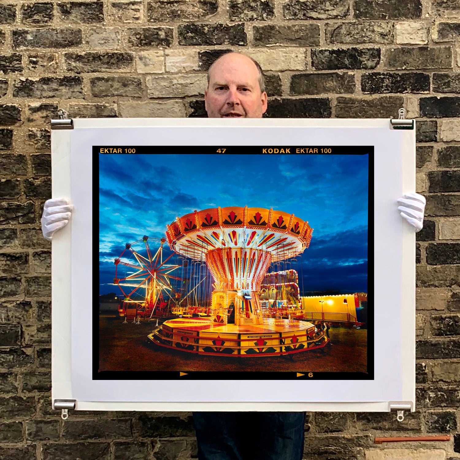 Photograph held by the photographer Richard Heeps. A fairground ride, the chairoplanes, sits lit in golden and red colours against a dark blue sky.
