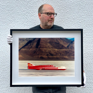 Photograph held by photographer Richard Heeps.  A Red Ferguson Racing Streamliner sits on a smooth salt flat with mountains in the background.