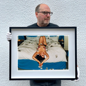 Black framed photograph held by photographer Richard Heeps. A woman lies semi naked on a bed with money surrounding her. The photograph is taken from the end of the bed so the woman appears upside down. The bed has a white duvet with blue under sheets.