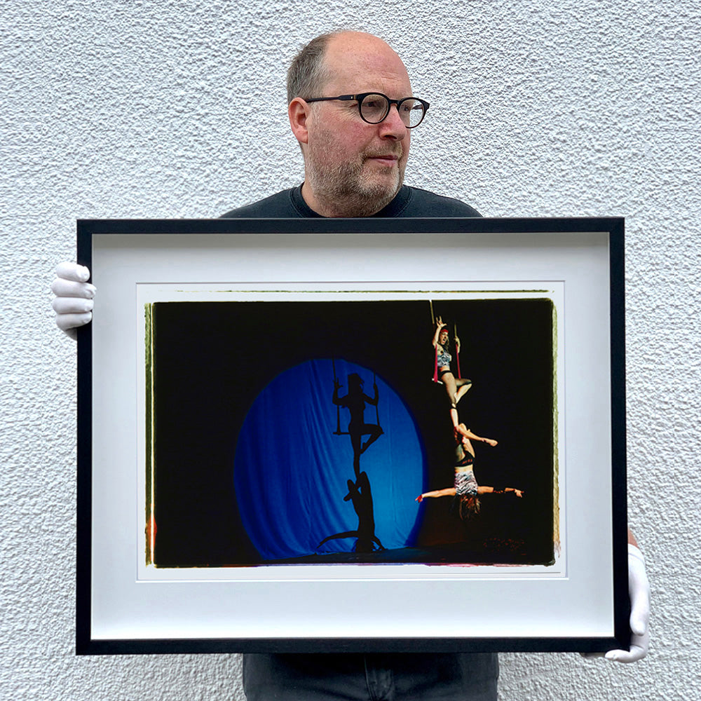 Black framed photograph held by photographer Richard Heeps. The photo is of two women on a trapeze, one sits on the swing and the other is hanging down from the first. The background is dark apart from a blue circle in which shows their acrobatic shadow.