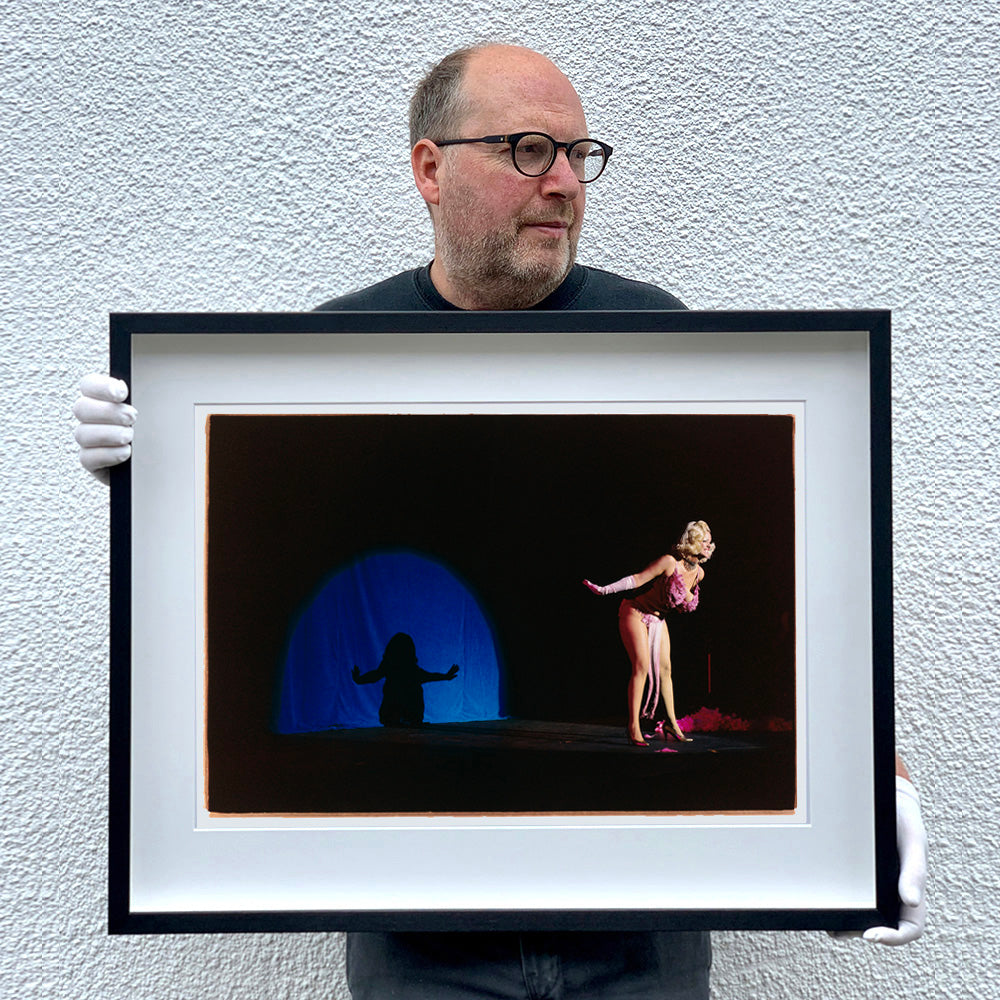 Photograh held by photographer Richard Heeps. A Burlesque dancer with a pink fluffy bikini bows on a black stage, in the background is a blue circle containing her silhouette.