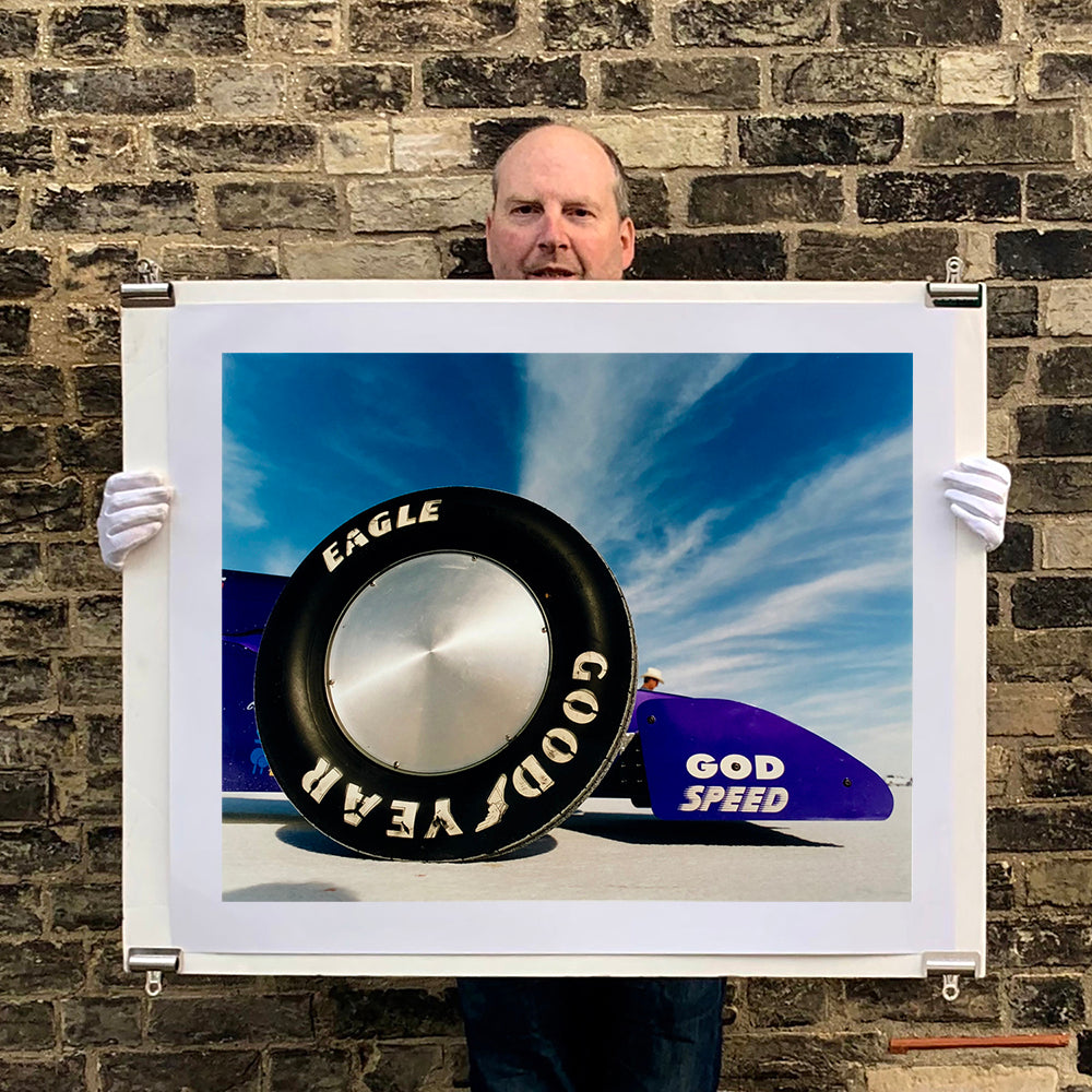 Photograph held by photographer Richard Heeps. This photograph has the tyre and the very front tip of a drag car. The car's name is written on the front end "God Speed". Behind the car are white vertical clouds shooting through a blue sky.