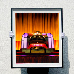 Framed artwork of a theater stage with 'STATE' on the podium against an orange curtain.