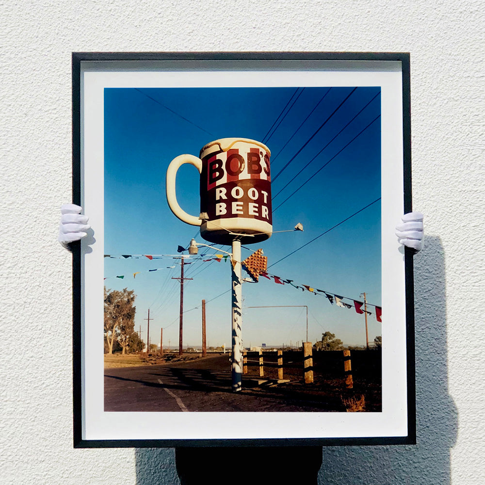 Photograph held by photographer Richard Heeps. A giant model of a mug with Bob's Root Beer written on it sits on top of a giant pole. There is bunting hanging from the pole. It sits alongside a power line on a remote looking American country road.