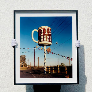Photograph held by photographer Richard Heeps. A giant model of a mug with Bob's Root Beer written on it sits on top of a giant pole. There is bunting hanging from the pole. It sits alongside a power line on a remote looking American country road.