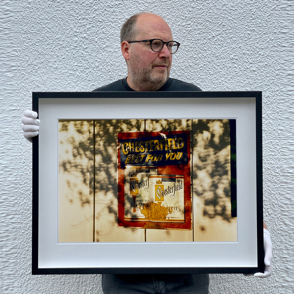 Black framed photograph held by photographer Richard Heeps. A plastic retro cigarette sign  for Chesterfield cigarettes is stuck to a white wash wooden fence. The words Chesterfield Best for You are raised on the sign and the sign is in a dappled light.