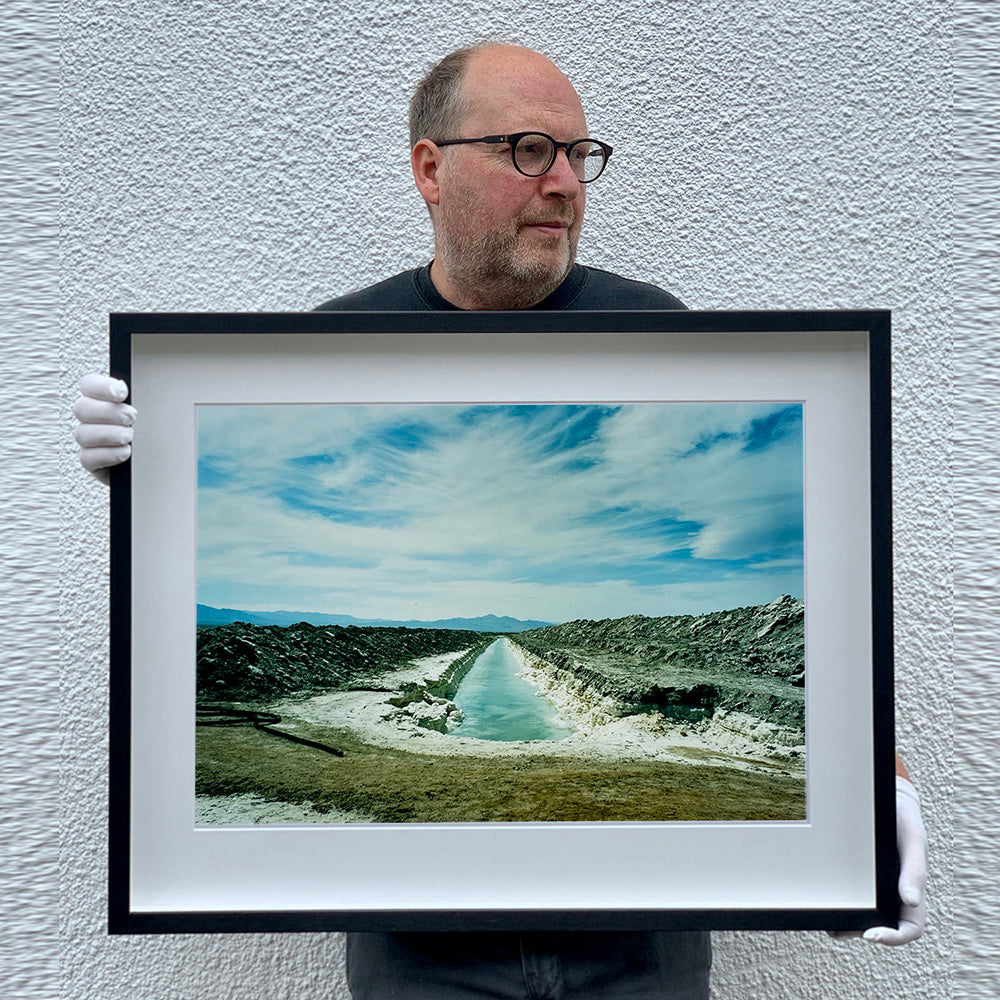 Black framed photograph held by photographer Richard Heeps. A watery white trench is dug into the centre going into the distance with grey mounds of earth either side. The sky is vast and blue, with light clouds.
