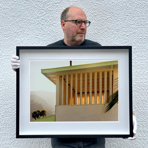 Black framed photograph held by photographer Richard Heeps.  The photograph is of a corner of the top of a cream colour building with big windows and vertical slats, on the left hand side are mountains bathing in the mist of dawn.