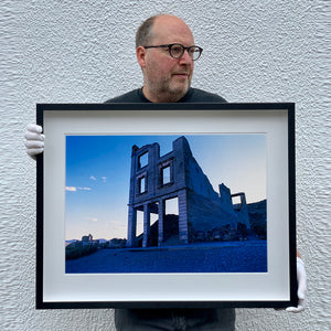 Black framed photograph held by photographer Richard Heeps. The remnants of a rectangular building sits alone, surrounded by rubble and gravel in a blue light.