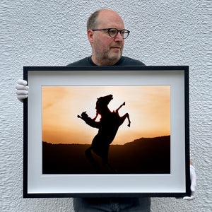 Black framed photograph held by Richard Heeps. The photograph shows a sunset and in the middle of the photograph is the silhouette of a statue of a cowboy on his bucking horse with one arm on the reins and the other held high.