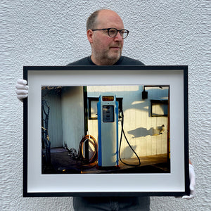 Black framed photograph held by Richard Heeps. A vintage petrol pump with a white front and blue sides, sitting outside a white slatted building.