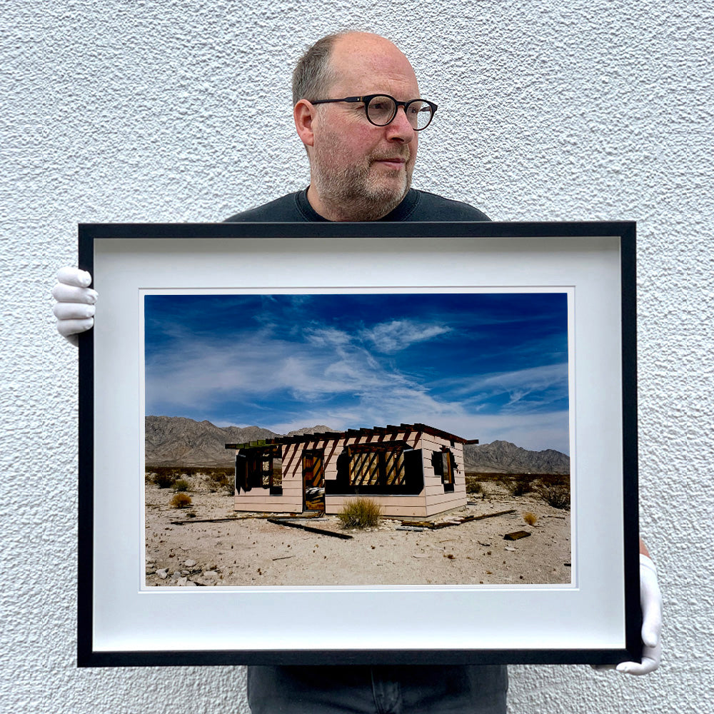 Photograph held by photographer Richard Heeps. An abandoned building sitting alone in a desert, tumble weed on the ground and hills in the background. A blue sky with white clouds.
