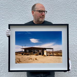 Photograph held by photographer Richard Heeps. An abandoned building sitting alone in a desert, tumble weed on the ground and hills in the background. A blue sky with white clouds.