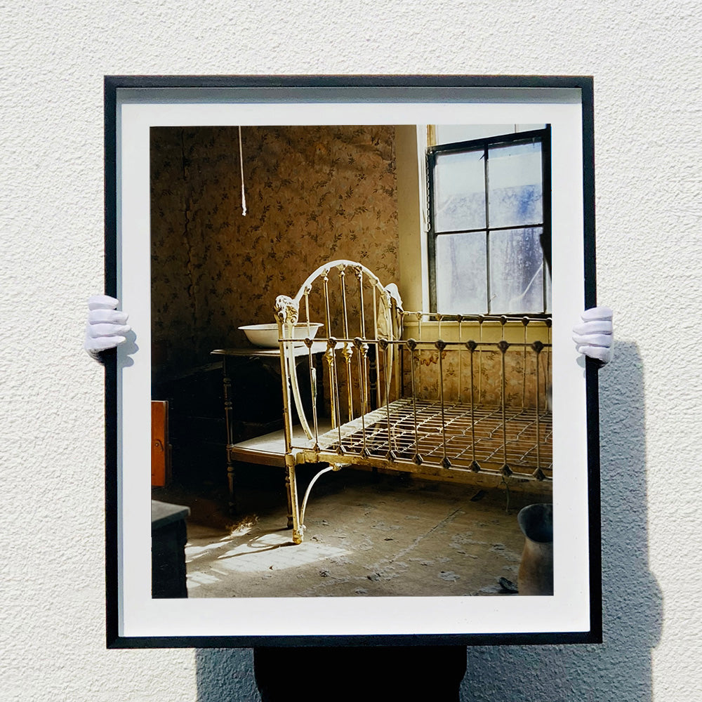Black photograph held by photographer Richard Heeps. A run down now unused room with the metal surround of a cot bed and no mattress, at the end of the bed a wash stand with a bowl on top. Light shines in the room from the window.