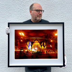 Black framed photograph held by photographer Richard Heeps. Photograph taken at night of the front of a brightly lit hotel. The scene is bathed in golden and reddish brown colours. The name of the hotel is La Concha and its name is written in white neon against a red and golden shell design. The photograph is taken through glass and there are reflections of cars as well as the room from which the photograph is taken.