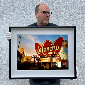 Black framed photograph held by Richard Heeps. This photograph is of the outside of La Concha Motel. The gold flamboyant La Concha lettering is set on a big red background. Below the motel sign is NO VACANCY with just VACANCY lit in red, below this sits a sign for Budget rent a car. Other signs and palm trees are the background together with a blue sky.