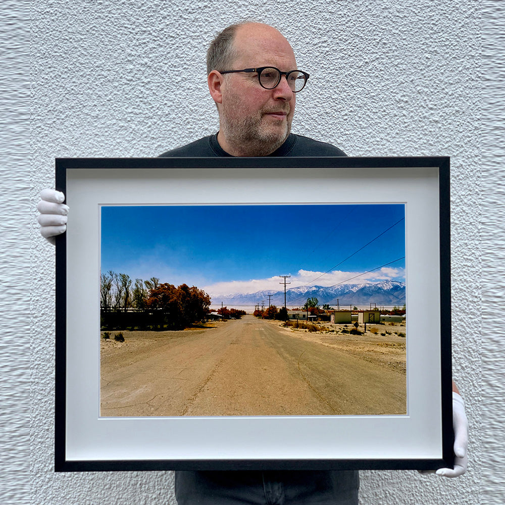 Black framed photograph held by Richard Heeps. A dusty road in the middle, heading towards the snow capped mountains in the distance, on the right are brown bushes and trees and on the left, single level concrete buildings.