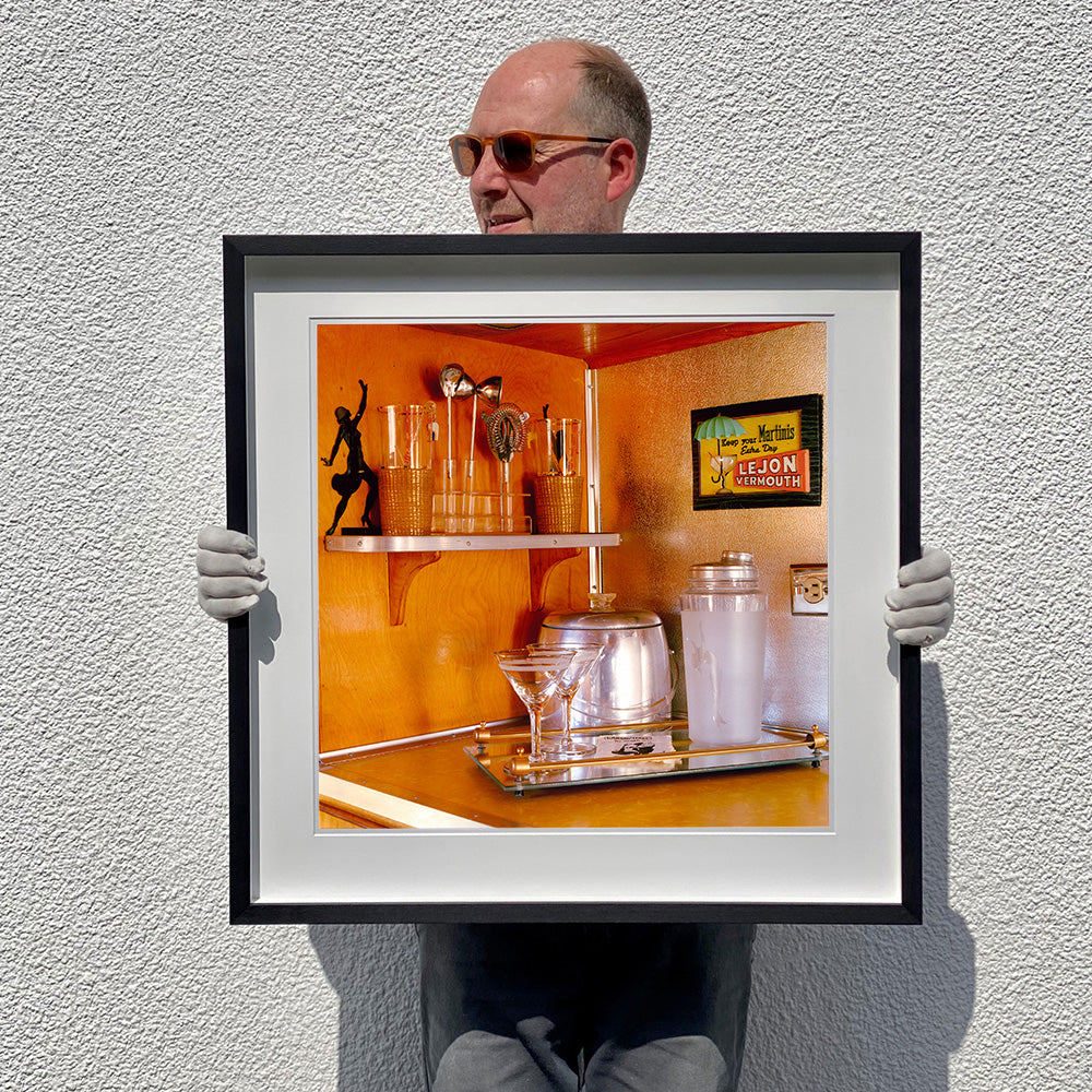 Black framed photograph held by photographer Richard Heeps. A drinks corner with martini glasses, cocktail shaker and ice bucket. There is a shelf with cocktail making equipment and it is set against retro orange walls.