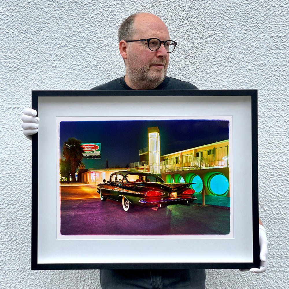 Black framed photograph by Richard Heeps. A Chevy Bel Air is central shot and off to the right are the pools and balcony of the Glass Pool Motel, Las Vegas. The photograph is being held by Richard Heeps.