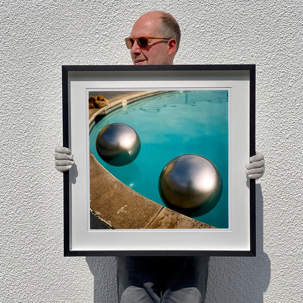 Black framed photograph held by photographer  Richard Heeps. The corner of a circular swimming pool with two metallic silver beach balls floating on the water.