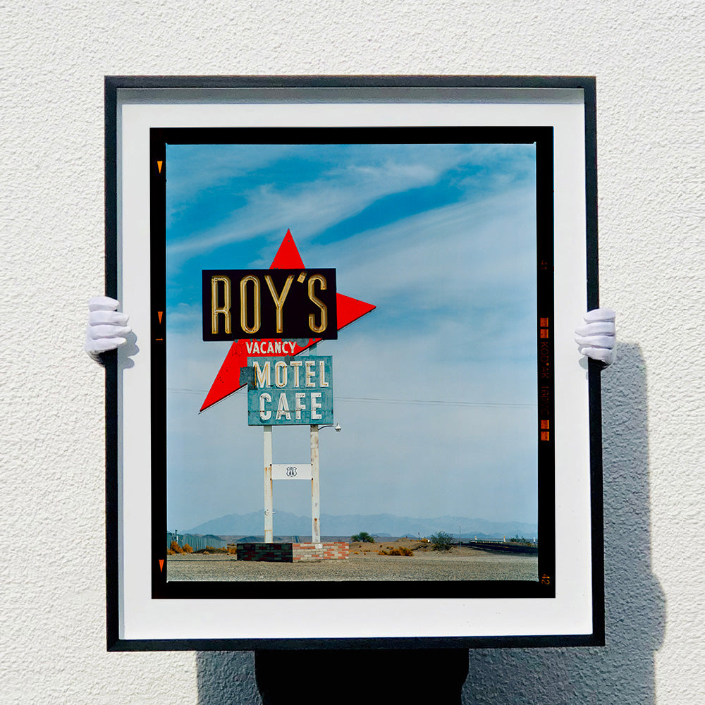 Photograph held by photographer Richard Heeps edged by film rebate. A roadside sign on Route 66 in America. The word ROY'S appears in a black sign with a bit red arrow pointing to the left ground, below this VACANCY and on a green square the words MOTEL and CAFE.