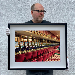 Photograph of a black framed photograph held by the photographer, Richard Heeps. The photograph depicts a line of slot machines sitting in a vintage Las Vegas casino. Perspective moves the slot machines from big machines on the left hand side to smaller on the right hand side. In front of the machines are a row of neatly lined blue stools one per machine and then red spectators chairs are neatly lined sitting forefront of the shot.