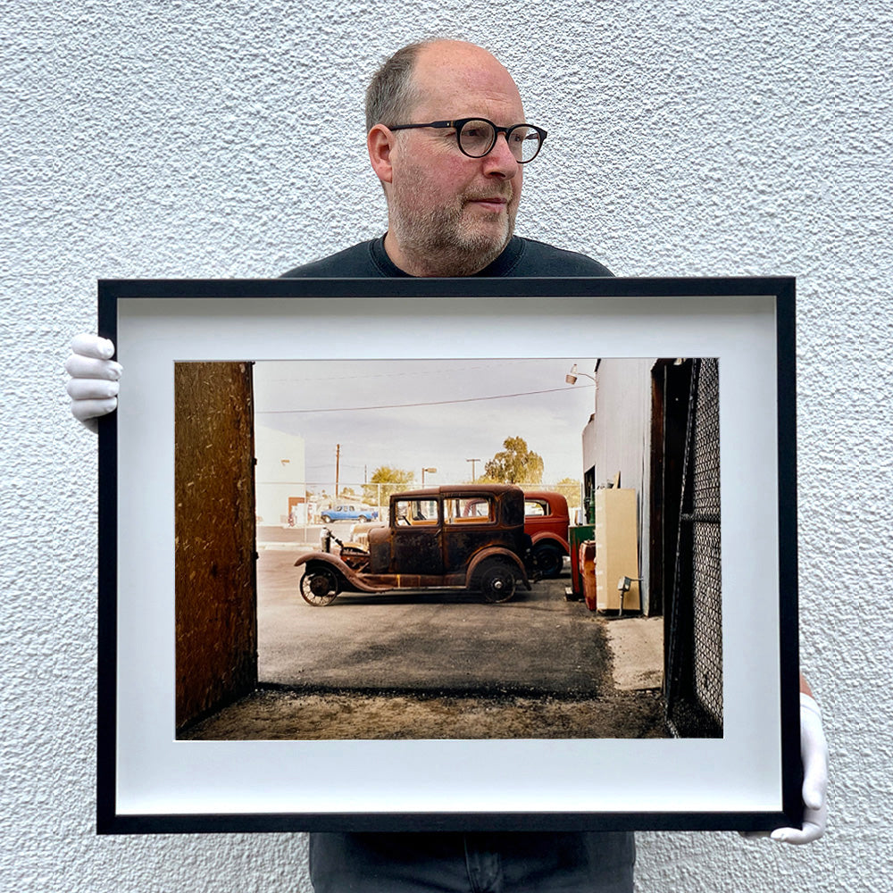 Black framed photograph held by Richard Heeps. The side view of two vintage early Ford Motor vehicles parked in a yard. 